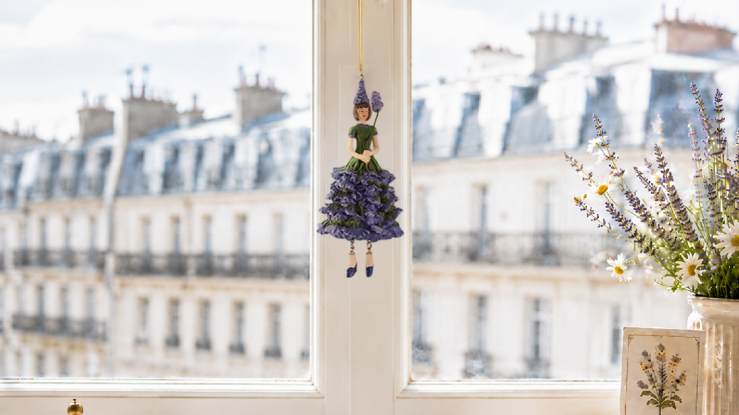 Figurine de petite fille aux fleurs bleue et verte suspendue à une fenêtre, devant les toits d’un immeuble parisien