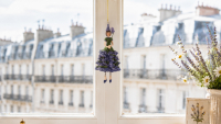 Figurine de petite fille aux fleurs bleue et verte suspendue à une fenêtre, devant les toits d’un immeuble parisien