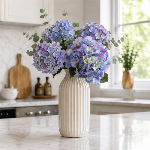 Blue artificial hydrangeas arranged in a tall ceramic vase in a bright kitchen