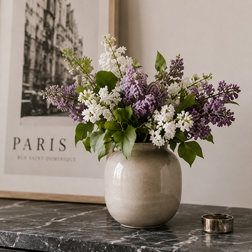 Weiße und lilafarbene Kunstblumen in einer graubeigen Vase auf dunklem Marmortisch